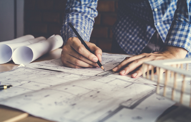 A man focuses on a blueprint, sketching details with a pencil at a desk.